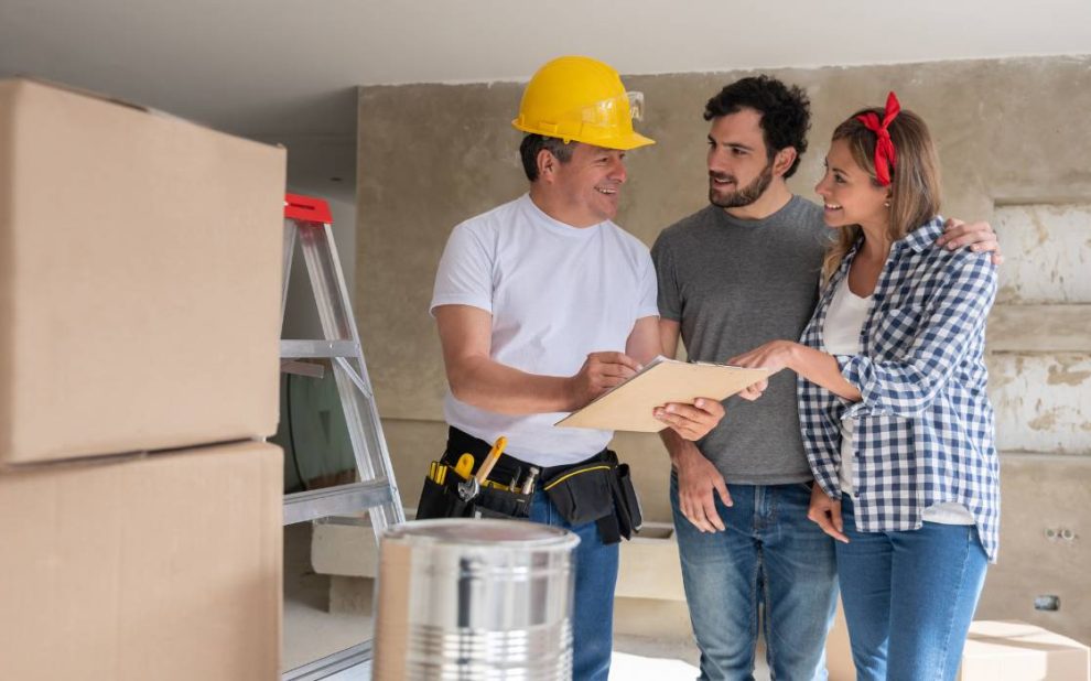 A home renovation contractor and a couple are standing in a room with boxes and a ladder.