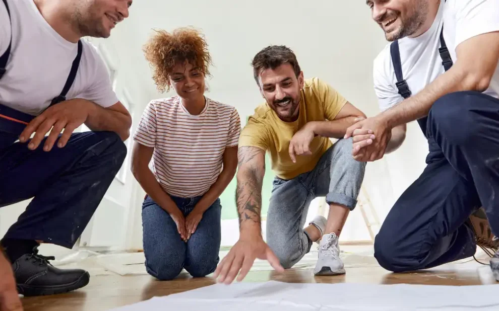 two home renovation contractors and a couple are looking at a blueprint on the floor while smiling.