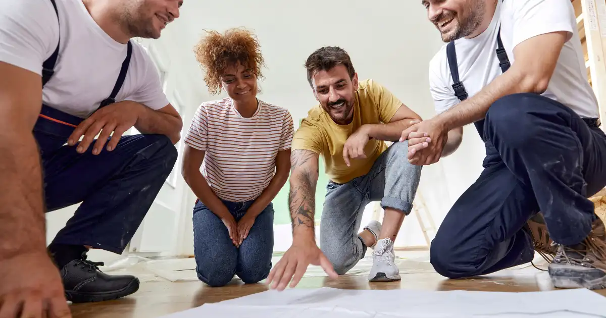 home-renovation-tips-1 two home renovation contractors and a couple are looking at a blueprint on the floor while smiling.